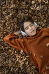 Boy with eyes closed listening to music through wireless headphones lying on dry leaves - ANAF00274