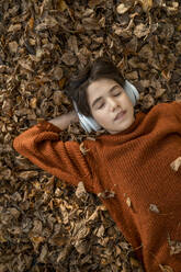 Junge mit geschlossenen Augen, der über kabellose Kopfhörer auf trockenem Laub liegend Musik hört - ANAF00274
