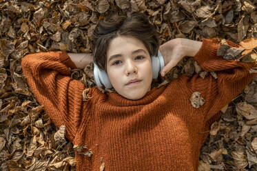 Junge hört Musik über kabellose Kopfhörer und liegt auf trockenem Laub - ANAF00275