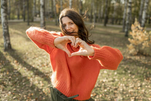 Smiling woman wearing orange sweater making heart shape with hands in park - ANAF00278