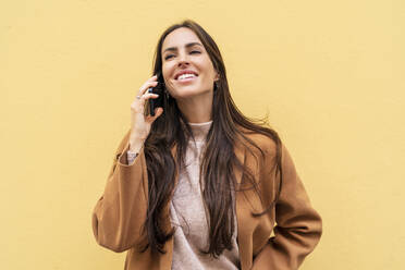 Glückliche junge Frau, die vor einer gelben Wand mit einem Mobiltelefon spricht - DLTSF03303