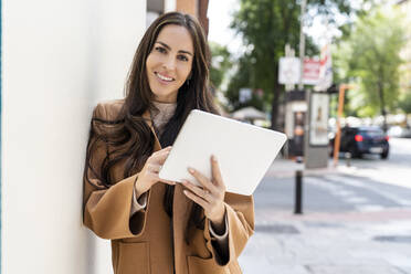 Smiling woman holding tablet PC leaning on white wall - DLTSF03307