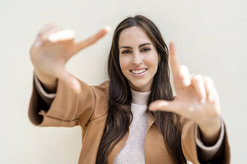 Smiling woman making picture frame with hands in front of white wall - DLTSF03308