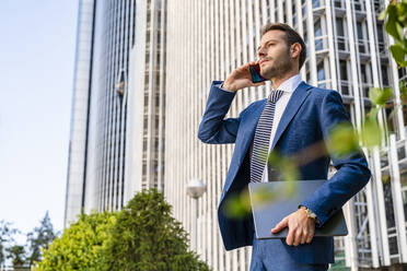 Businessman talking over mobile phone holding laptop at office park - DLTSF03318