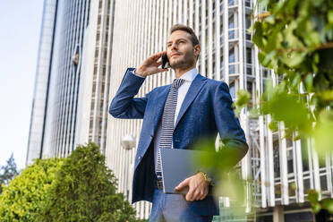 Businessman talking through mobile phone holding laptop at office park - DLTSF03319