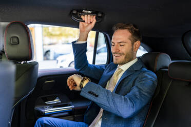 Happy businessman checking time sitting on back seat in taxi - DLTSF03329