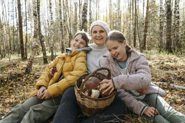 Smiling grandmother, boy and girl sitting with basket of mushrooms in forest - EYAF02271