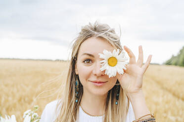 Lächelnde Frau, die ihr Auge mit einer weißen Blume im Feld bedeckt - MDOF00143