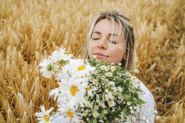 Lächelnde Frau mit geschlossenen Augen, die einen Strauß Gänseblümchen auf einem Feld hält - MDOF00144
