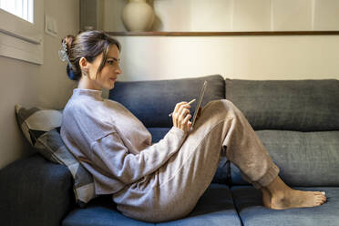 Young woman using tablet PC sitting on sofa in living room at home - DLTSF03332