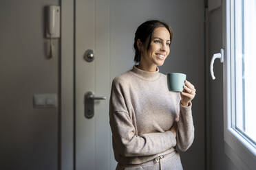 Glückliche Frau mit Kaffeetasse am Fenster stehend zu Hause - DLTSF03334