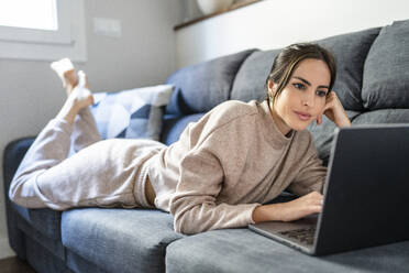 Junge Frau, die auf dem Sofa liegend einen Film über einem Laptop ansieht - DLTSF03335