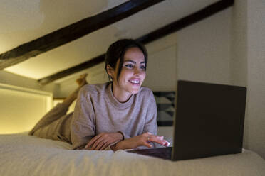 Smiling young woman using laptop on bed in bedroom - DLTSF03336