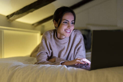 Glückliche junge Frau mit Laptop auf dem Bett liegend zu Hause - DLTSF03337