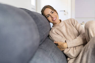 Glückliche junge Frau sitzt auf dem Sofa im Wohnzimmer - DLTSF03338