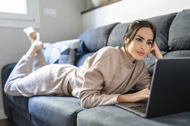 Junge Frau sieht sich einen Film auf dem Laptop an und liegt zu Hause auf der Couch - DLTSF03345