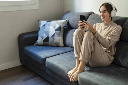 Frau mit Smartphone und Kopfhörern beim Musikhören auf dem Sofa zu Hause - DLTSF03346