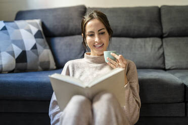 Junge Frau liest ein Buch mit einer Tasse Kaffee zu Hause - DLTSF03347