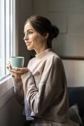 Thoughtful woman standing with cup of tea near window at home - DLTSF03348
