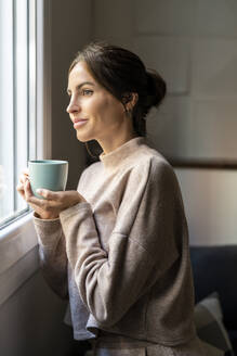 Nachdenkliche Frau steht mit einer Tasse Tee in der Nähe des Fensters zu Hause - DLTSF03348