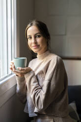 Smiling woman with cup of tea standing by window at home - DLTSF03349