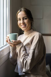 Happy young woman with cup of tea standing by window at home - DLTSF03350