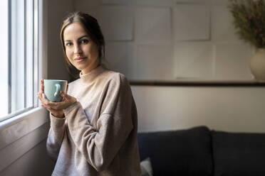 Lächelnde junge Frau mit einer Tasse Tee am Fenster zu Hause - DLTSF03351