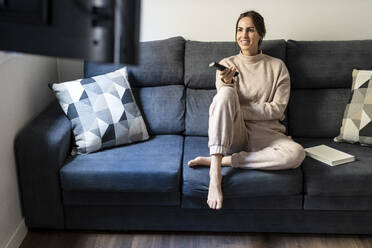 Smiling woman holding remote control sitting on sofa at home - DLTSF03352