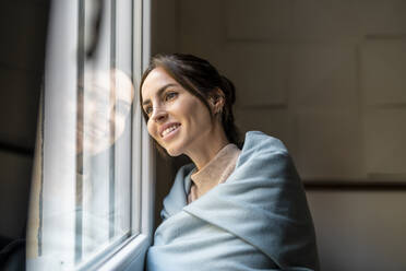 Nachdenkliche junge Frau in eine Decke gewickelt am Fenster stehend - DLTSF03356