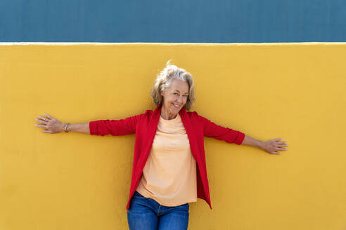 Lächelnde Frau mit ausgestreckten Armen, die sich an eine gelbe Wand lehnt - OIPF02460