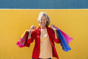 Happy mature woman with multi colored shopping bags in front of yellow wall - OIPF02466