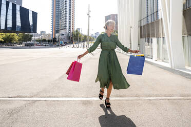 Cheerful mature woman carrying shopping bags and walking on footpath in city - OIPF02491