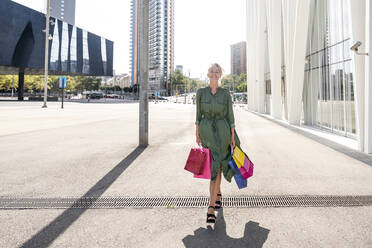 Smiling woman with shopping bags walking on footpath in city - OIPF02494