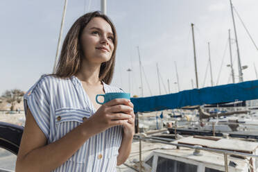 Smiling young woman with coffee cup standing at harbor - JRVF03151
