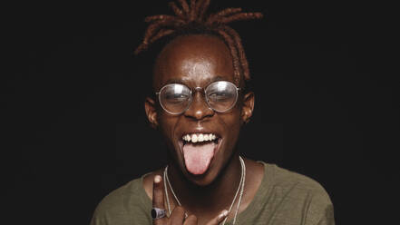 Afrikanischer Mann mit Dreadlocks, der seine Zunge herausstreckt und in die Kamera schaut. Nahaufnahme eines lachenden afrikanischen Mannes, der ein lustiges Gesicht macht, isoliert auf schwarzem Hintergrund. - JLPSF17798