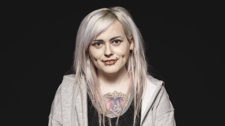 Smiling woman with white hair and tattoo on body looking at camera. Woman with fashion piercing on lips and cheeks isolated on black background. - JLPSF17994