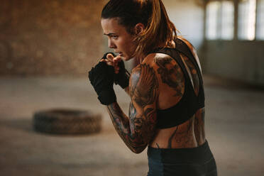 Female doing shadow boxing in empty factory shade. Tattooed woman in sportswear practicing her punches at an old warehouse. - JLPSF18007