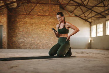 Fitness-Frau, die sich nach dem Training ausruht und ein Mobiltelefon benutzt. Frau sitzt auf einer Trainingsmatte und benutzt ein Mobiltelefon in einem verlassenen Lagerhaus. - JLPSF18010