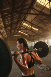 Fit woman exercising using barbell with heavy weights in a gym inside empty warehouse. Female doing squats exercise. - JLPSF18018