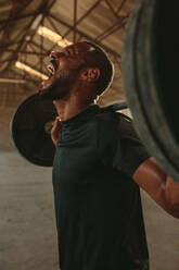 Ein Mann schreit, während er mit schweren Gewichten im Fitnessstudio trainiert. Ein starker Mann trainiert mit einer Langhantel. Er strengt sich an, während er in einem alten Lagerhaus trainiert. - JLPSF18020