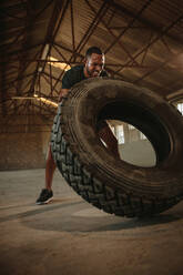 Ein starker Mann wirft einen Reifen während einer intensiven Trainingseinheit. Ein fitter Mann macht ein Reifenflipping-Training in einem leeren Lagerhaus. - JLPSF18021