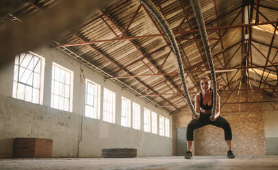 Starke Frau, die ein Paar Kampfseile für Übungen in einem Fitnessstudio im Schatten einer alten Fabrik benutzt. - JLPSF18028