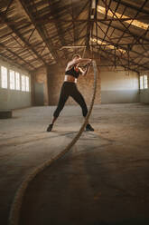 Strong woman using training ropes for exercise. Female working out with battle rope at gym inside old warehouse - JLPSF18029