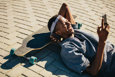 Lächelnder afroamerikanischer Mann, der auf seinem Skateboard liegt und Textnachrichten auf seinem Mobiltelefon liest. Skateboarder, der in einer Pause sein Smartphone benutzt. - JLPSF18038