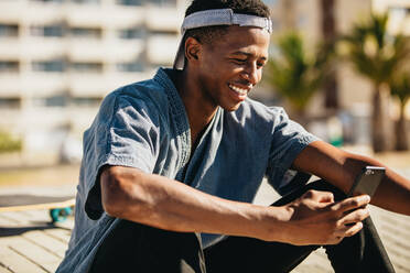 Lächelnder afroamerikanischer Mann, der im Freien sitzt und Textnachrichten auf seinem Smartphone liest. Junger Skateboarder, der lächelt, während er ein Mobiltelefon benutzt. - JLPSF18039