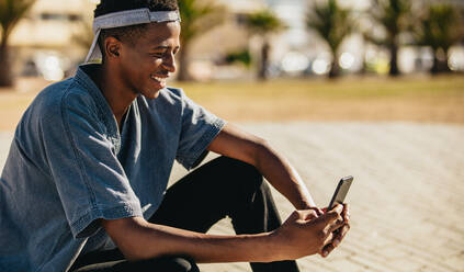 Afroamerikanischer junger Mann sitzt im Freien und benutzt ein Handy. Junger Mann schreibt eine SMS mit seinem Handy und lächelt, - JLPSF18040