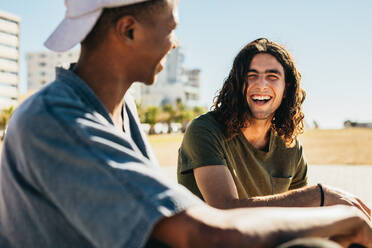 Junger Mann lächelt im Gespräch mit seinem Freund im Freien. Zwei junge Männer sitzen im Freien und lächeln sich an. - JLPSF18041
