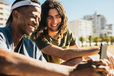 Junger Mann und sein Freund schauen auf ein Handy und lächeln im Freien. Zwei junge Männer sitzen im Freien und benutzen ein Smartphone. - JLPSF18042
