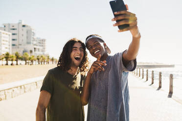 Zwei junge, gemischtrassige Männer machen ein Selfie an der Strandpromenade. Glückliche junge Freunde, die ein Selbstporträt mit einem Smartphone im Freien machen. - JLPSF18045