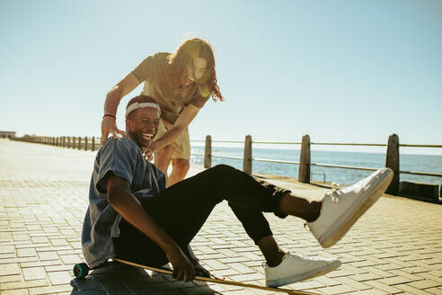 Junger Mann, der seinen Freund auf einem Longboard an der Strandpromenade schiebt. Jungen, die sich mit einem Longboard auf einer Straße am Meer vergnügen. - JLPSF18049